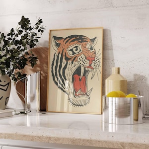 May include: Framed vintage-style tiger art print. The tiger's head is depicted with orange, black, and red colors, with its mouth open in a roar. The artwork is in a light-colored wooden frame, displayed on a white countertop.