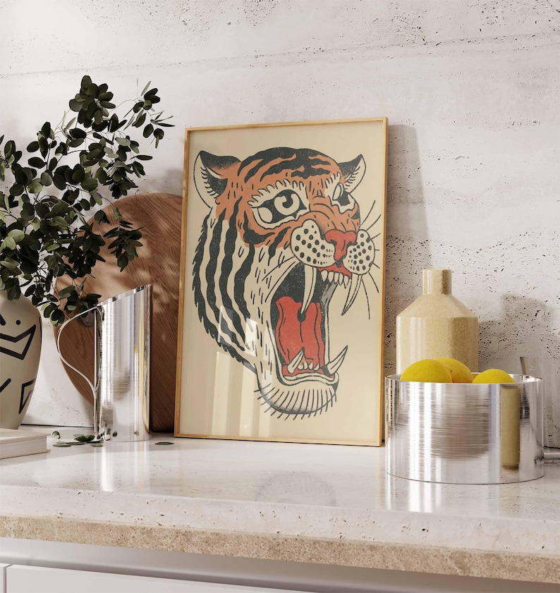 May include: Framed vintage-style tiger art print. The tiger's head is depicted with orange, black, and red colors, with its mouth open in a roar. The artwork is in a light-colored wooden frame, displayed on a white countertop.