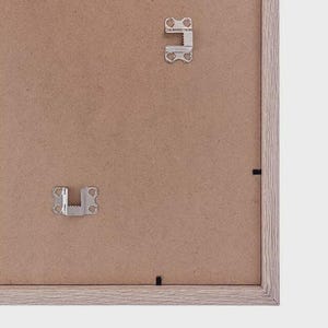 May include: The reverse side of a picture frame, showing metal hanging brackets and a light brown cardboard backing. The frame itself has a light brown, wood-effect finish. The frame is on a white background. The metal brackets are for hanging the picture.