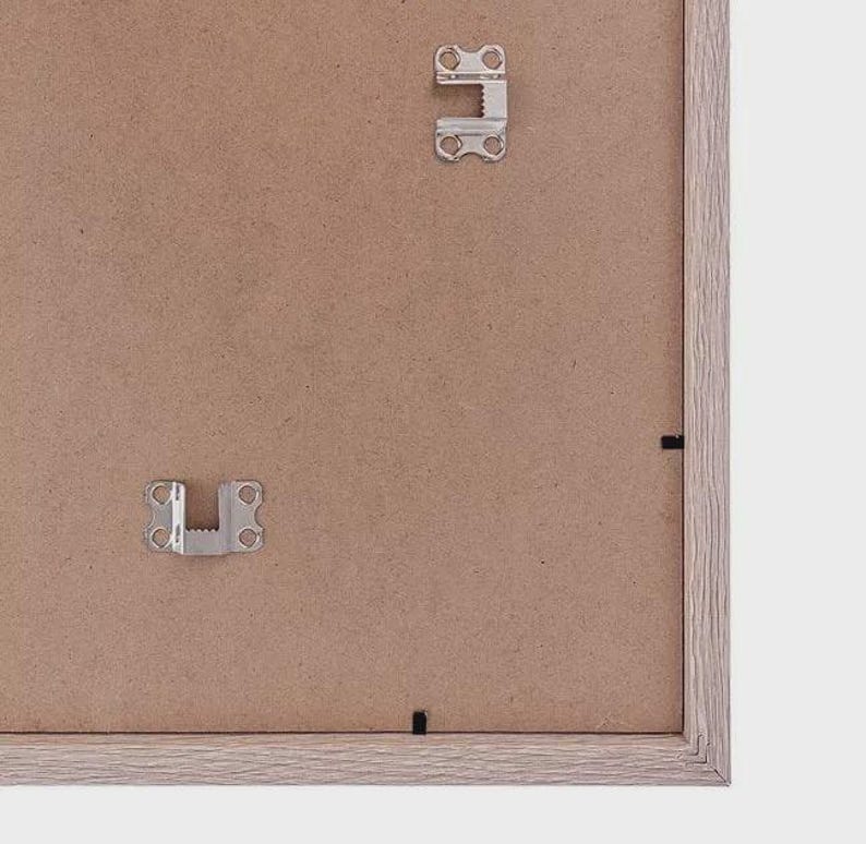 May include: The reverse side of a picture frame, showing metal hanging brackets and a light brown cardboard backing. The frame itself has a light brown, wood-effect finish. The frame is on a white background. The metal brackets are for hanging the picture.