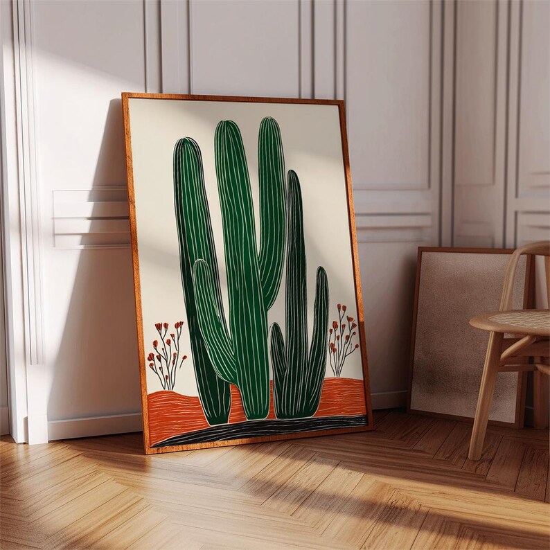 May include: A framed art print featuring a stylized illustration of green cacti against a cream background. The cacti are set on an orange and black ground, with small red floral accents. The frame is a warm brown color.