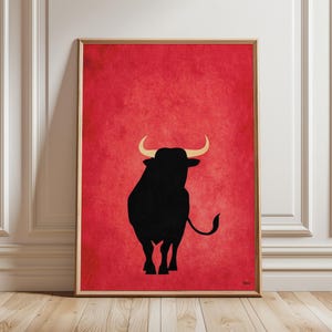 Può includere: Stampa artistica incorniciata con una silhouette nera di un toro su uno sfondo rosso strutturato. Il toro ha corna color oro. L'opera d'arte è esposta in una stanza con modanature bianche e pavimento in legno.