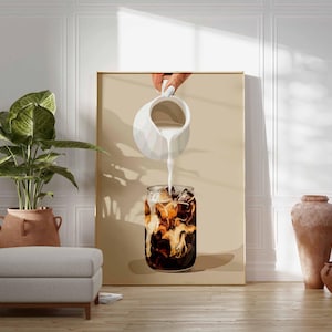 May include: A framed art print depicts a refreshing iced coffee. A hand pours cream from a white pitcher into a glass filled with ice and dark coffee. The artwork is set against a neutral background, with a plant, ottoman, and pottery in the foreground.