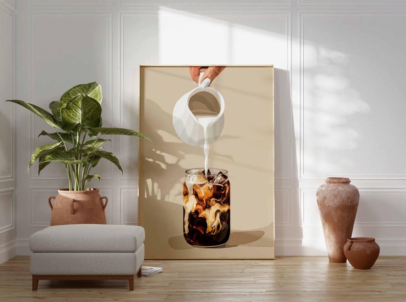 May include: A framed art print depicts a refreshing iced coffee. A hand pours cream from a white pitcher into a glass filled with ice and dark coffee. The artwork is set against a neutral background, with a plant, ottoman, and pottery in the foreground.