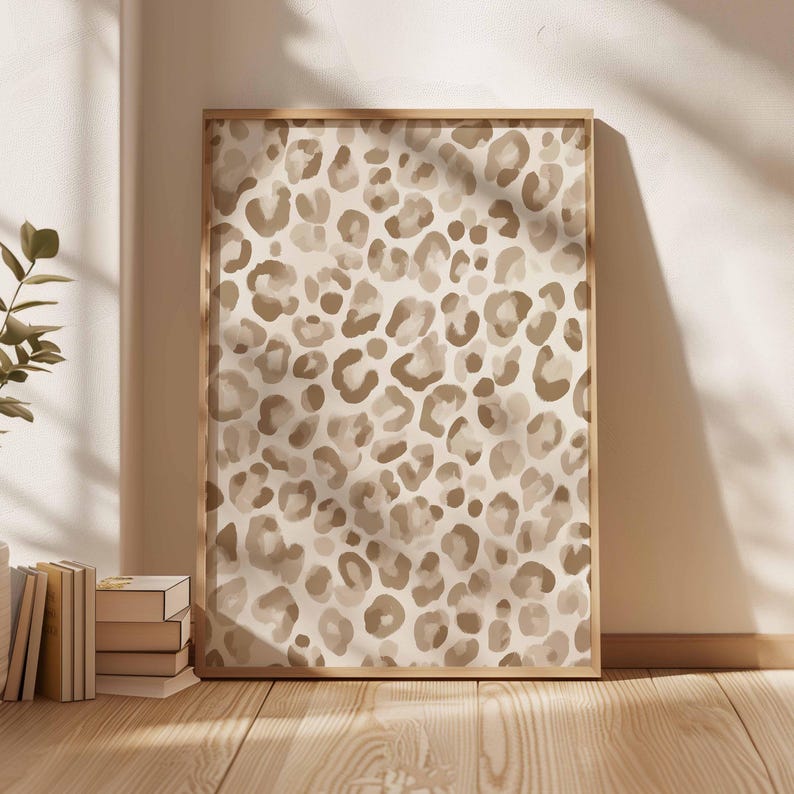 May include: A framed art print featuring a leopard print pattern in shades of brown and beige. The artwork is displayed in a light-colored wooden frame and leans against a white wall. The print's design is a repeating pattern of irregular spots.