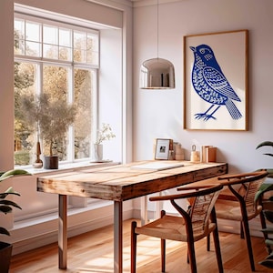 May include: A framed print featuring a blue bird design hangs above a wooden desk with metal legs. Two woven chairs are positioned in front of the desk. A large window offers a view of trees and natural light. A silver pendant light hangs above the desk.