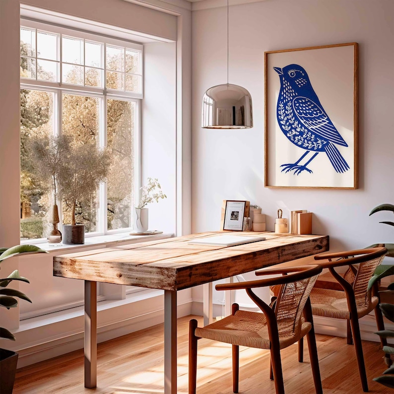 May include: A framed print featuring a blue bird design hangs above a wooden desk with metal legs. Two woven chairs are positioned in front of the desk. A large window offers a view of trees and natural light. A silver pendant light hangs above the desk.