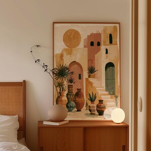 Peut inclure: Une impression d'art encadr&eacute;e repr&eacute;sente un paysage urbain abstrait aux tons chauds, avec des arches, des escaliers et un grand soleil. L'&oelig;uvre est expos&eacute;e au-dessus d'un meuble en bois avec un vase rond, des livres et une lampe globe. Un cadre de lit est visible &agrave; gauche.