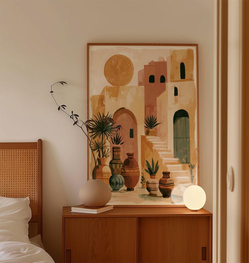 Peut inclure: Une impression d'art encadr&eacute;e repr&eacute;sente un paysage urbain abstrait aux tons chauds, avec des arches, des escaliers et un grand soleil. L'&oelig;uvre est expos&eacute;e au-dessus d'un meuble en bois avec un vase rond, des livres et une lampe globe. Un cadre de lit est visible &agrave; gauche.