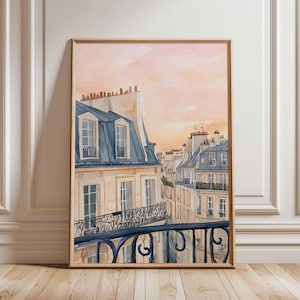 May include: Watercolour painting of Parisian buildings with blue rooftops and a wrought iron balcony. The artwork is framed in a light brown wooden frame. The sky is a soft pink and orange, suggesting a sunset or sunrise.