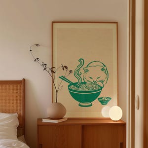 May include: A framed wall art print featuring a green illustration of a cat eating noodles from a bowl with chopsticks. The print is set against a light beige background and is displayed in a simple wooden frame on a wooden cabinet.