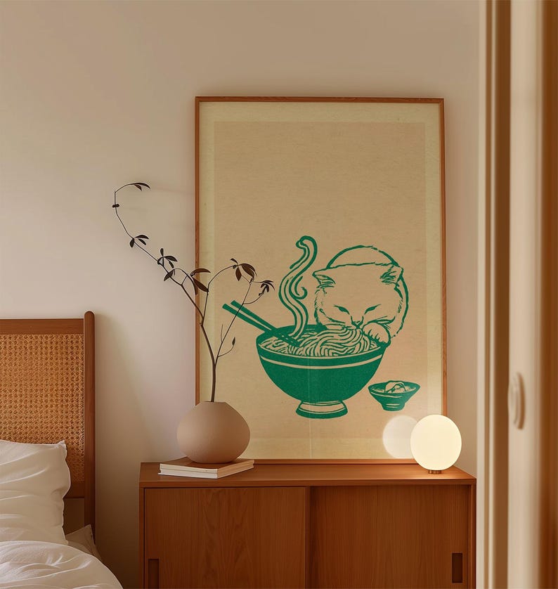 May include: A framed wall art print featuring a green illustration of a cat eating noodles from a bowl with chopsticks. The print is set against a light beige background and is displayed in a simple wooden frame on a wooden cabinet.