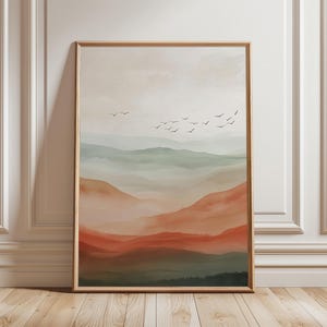 May include: A framed landscape art print with a watercolour painting of layered mountains in shades of green, orange, and brown. A flock of birds flies across the soft, neutral-toned sky. The artwork is encased in a light brown wooden frame.