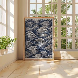 May include: A framed art print featuring a navy blue and white wave pattern. The artwork is displayed in a light wooden frame and leans against a wall in a room with a large window and hardwood floors. Potted plants sit on the windowsill.