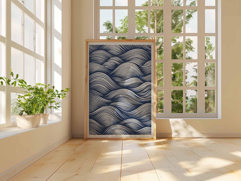 May include: A framed art print featuring a navy blue and white wave pattern. The artwork is displayed in a light wooden frame and leans against a wall in a room with a large window and hardwood floors. Potted plants sit on the windowsill.