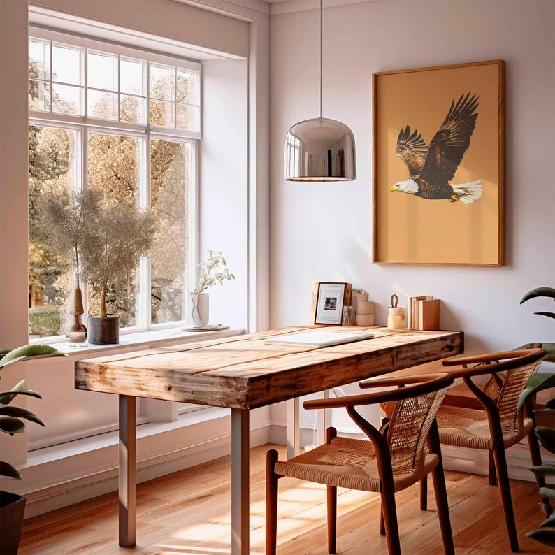 May include: A rustic wooden desk with metal legs and two woven chairs sits in a bright room. A framed eagle print hangs on the wall. A large window offers a view of trees. A silver pendant light hangs above the desk.