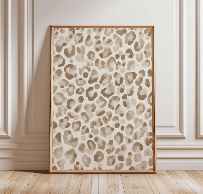 May include: A framed art print featuring a beige and brown leopard print pattern. The artwork is displayed in a light brown wooden frame and leans against a white wall and wooden floor.