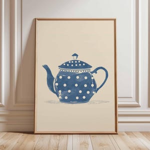 Op de afbeelding: Een ingelijste print met een blauwe theepot met witte stippen. De theepot heeft een gebogen tuit en handvat, en een deksel met een kleine knop. De achtergrond is lichtbeige en de lijst is van natuurlijk hout.