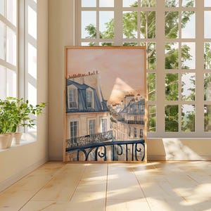 May include: A framed watercolour print of a Parisian street scene. The artwork depicts buildings with blue-grey roofs and wrought-iron balconies, set against a soft pink and orange sky. The print is displayed in a light-coloured wooden frame.