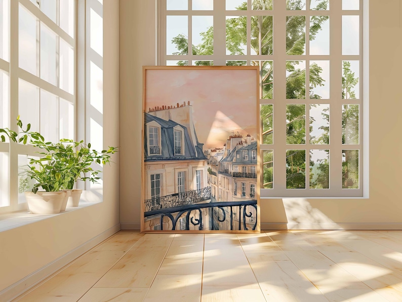 May include: A framed watercolour print of a Parisian street scene. The artwork depicts buildings with blue-grey roofs and wrought-iron balconies, set against a soft pink and orange sky. The print is displayed in a light-coloured wooden frame.