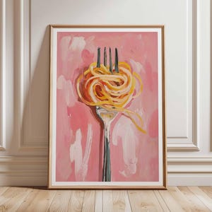 Op de afbeelding: Een ingelijste kunstdruk met een vork die een werveling van spaghetti vasthoudt. De pasta is geschilderd in gele en oranje tinten, tegen een roze achtergrond met witte penseelstreken. De vork is zilverkleurig en het kunstwerk zit in een gouden lijst.