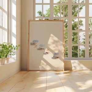 May include: A framed print showing a beach scene with parasols and people sunbathing. The artwork is displayed in a room with natural light from a large window, and a potted plant on the windowsill. The floor is wooden.