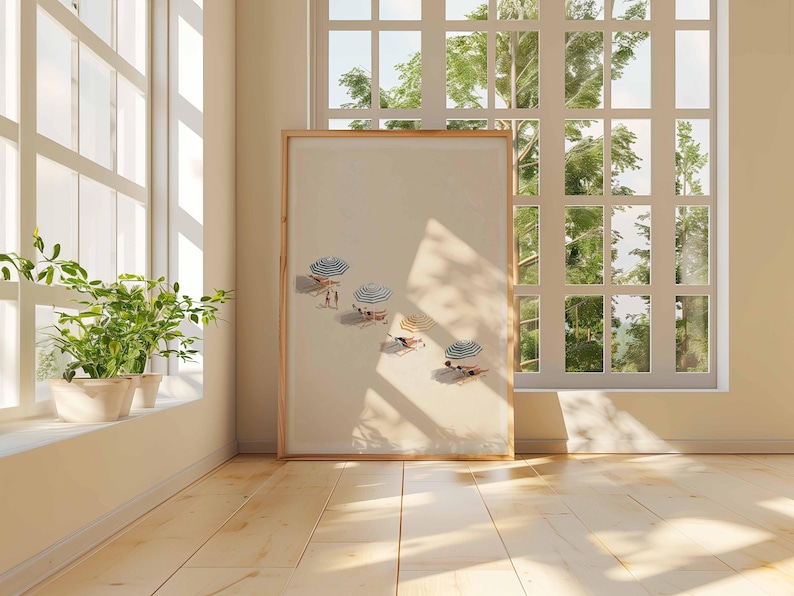 May include: A framed print showing a beach scene with parasols and people sunbathing. The artwork is displayed in a room with natural light from a large window, and a potted plant on the windowsill. The floor is wooden.