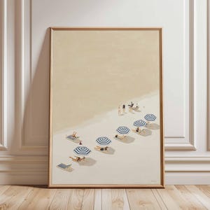 May include: A framed art print showing a beach scene with people relaxing under striped umbrellas. The artwork features a sandy beach, blue and white striped parasols, and sun loungers. The colour palette includes beige, blue, and brown tones.