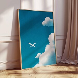 Peut inclure: Une affiche encadr&eacute;e repr&eacute;sentant un ciel bleu avec des nuages blancs et un petit avion blanc. L'&oelig;uvre d'art a un cadre en bois clair et est appuy&eacute;e contre un mur blanc et un sol en bois.