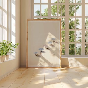 May include: A framed art print showing a beach scene with figures under striped umbrellas. The artwork is displayed in a room with a large window, letting in natural light. The print has a light wood frame.