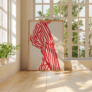 Puede incluir: Una obra de arte enmarcada que presenta una figura con un atuendo a rayas rojas y blancas. La obra de arte se exhibe en una habitaci&oacute;n con luz natural, piso de madera y una ventana con vistas a la vegetaci&oacute;n. La obra es el punto focal.