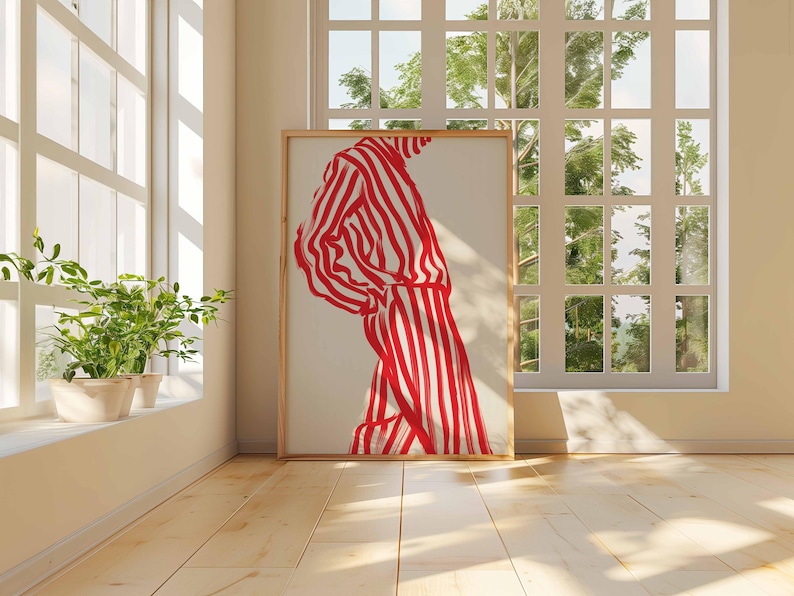Puede incluir: Una obra de arte enmarcada que presenta una figura con un atuendo a rayas rojas y blancas. La obra de arte se exhibe en una habitaci&oacute;n con luz natural, piso de madera y una ventana con vistas a la vegetaci&oacute;n. La obra es el punto focal.