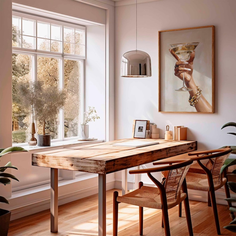 Op de afbeelding: Een lichte kamer met een houten bureau en twee stoelen. Een ingelijst schilderij van een hand die een cocktail vasthoudt, hangt aan de muur. Een groot raam biedt uitzicht op bomen. Een zilveren hanglamp hangt boven het bureau.