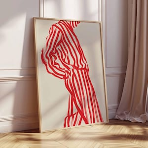 Puede incluir: Una impresi&oacute;n art&iacute;stica enmarcada que presenta una figura con un atuendo a rayas rojas y blancas. La obra de arte es una ilustraci&oacute;n minimalista con trazos audaces, sobre un fondo crema. El marco es de madera clara y la obra se apoya en una pared blanca.