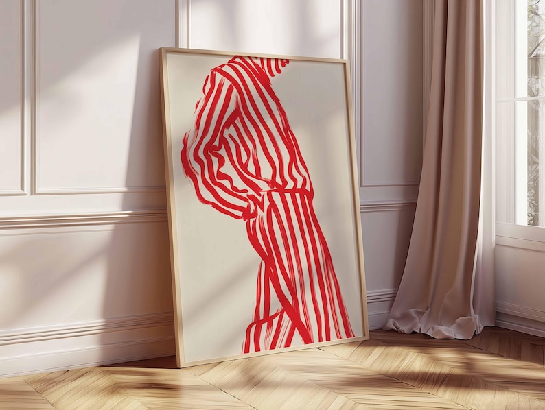 Puede incluir: Una impresi&oacute;n art&iacute;stica enmarcada que presenta una figura con un atuendo a rayas rojas y blancas. La obra de arte es una ilustraci&oacute;n minimalista con trazos audaces, sobre un fondo crema. El marco es de madera clara y la obra se apoya en una pared blanca.