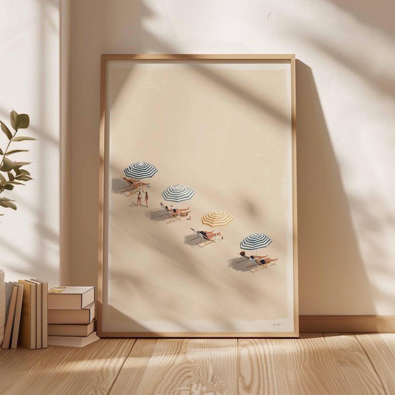 May include: A framed art print showing a beach scene with people relaxing under striped parasols. The artwork features a sandy beach with several deckchairs and parasols in shades of blue, yellow, and white. The print is displayed in a light-coloured wooden frame.