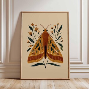 Può includere: Una stampa artistica incorniciata con un'illustrazione stilizzata di una falena. La falena ha ali arancioni e gialle con dettagli verde scuro, su uno sfondo beige. L'opera include elementi floreali ed è incorniciata in legno marrone chiaro.