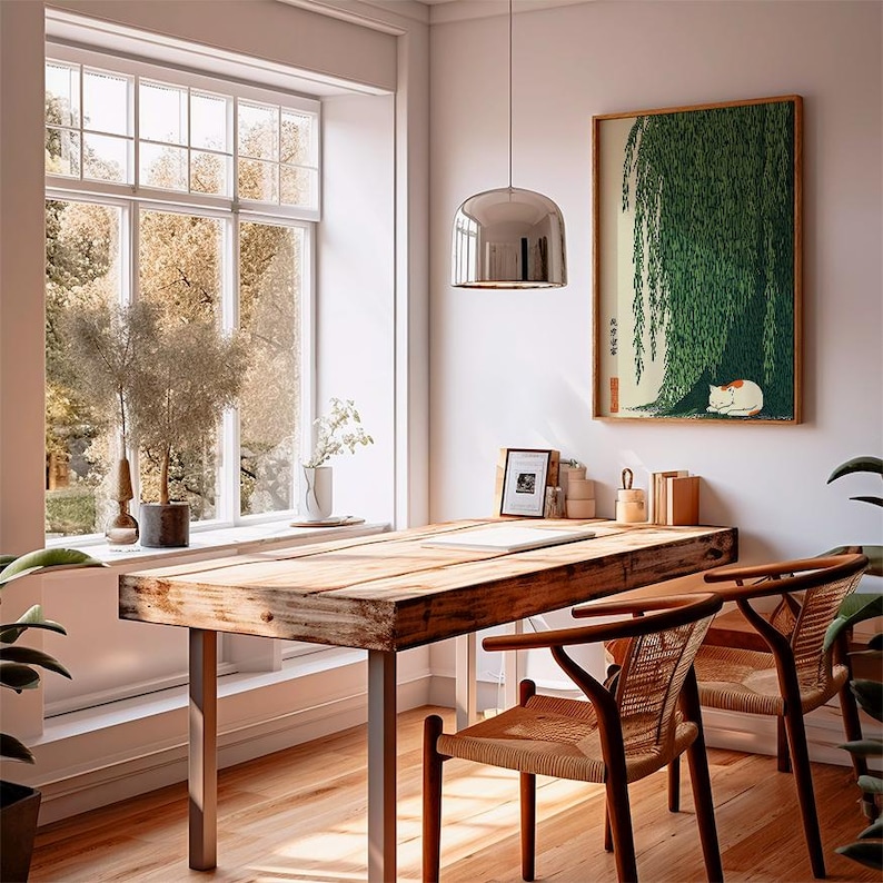 May include: A sunlit room with a large wooden desk and two woven chairs. A framed print of a cat sleeping under a weeping willow hangs on the wall. A large window offers a view of trees and natural light.