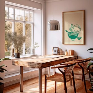 May include: A framed wall art print featuring a teal-colored illustration of a cat eating noodles from a bowl with chopsticks. The print is displayed on a light-colored wall above a wooden table and chairs in a bright, sunlit room.