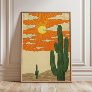 May include: A framed art print showing a desert scene with an orange sky and a yellow sun. Two green cacti are in the foreground, with a smaller one further away. The artwork creates a warm, sunny atmosphere, reminiscent of the American Southwest.