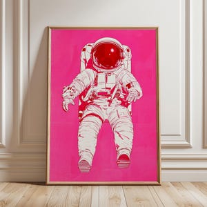 以下が含まれることがあります： 鮮やかなピンク色の背景に、白と赤の宇宙飛行士のイラストが描かれた額入りのアートプリント。宇宙飛行士は宇宙服とヘルメットを着用して座っています。アートワークは金色のフレームに収められています。