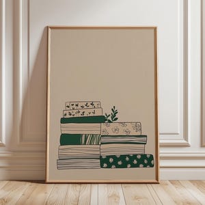 May include: A framed art print depicting a stack of books in green, beige, and white. The books display diverse patterns, including floral and striped designs. A sprig of leaves sits atop the stack. The background is a neutral beige colour.