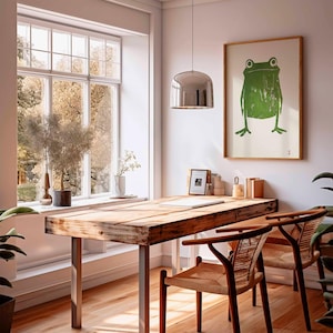 May include: A wooden table with a metal frame and two chairs. A framed print of a green frog is hanging on the wall above the table. A window with a view of trees is in the background.