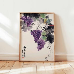 Op de afbeelding: Een ingelijste aquarel schildering van paarse druiven die aan wijnranken met groene bladeren hangen. Het kunstwerk heeft een traditionele Aziatische stijl, met kalligrafie en een rood zegel. De lijst is gemaakt van lichtgekleurd hout.