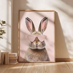May include: A framed print featuring a brown rabbit wearing pink heart-shaped sunglasses. The rabbit is centered against a soft pink background. The artwork is in a wooden frame, creating a whimsical and stylish wall decor piece.