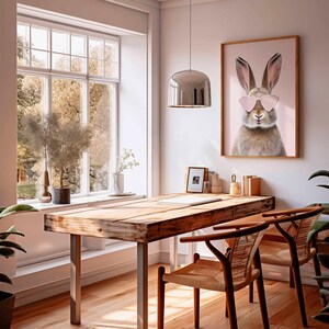 May include: A home office with a wooden desk and two chairs. A framed print of a rabbit wearing heart-shaped sunglasses hangs on the wall. A large window provides natural light, and a silver pendant light hangs above the desk.