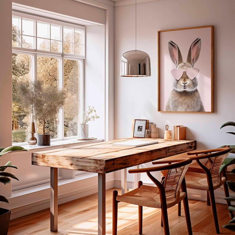 May include: A home office with a wooden desk and two chairs. A framed print of a rabbit wearing heart-shaped sunglasses hangs on the wall. A large window provides natural light, and a silver pendant light hangs above the desk.