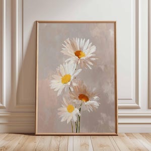 May include: A framed print of four white daisy flowers with yellow centres. The daisies are set against a soft, neutral background with a light brown wooden frame. The artwork is displayed on a wooden floor.