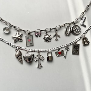 Könnte beinhalten: Zwei silberne Ketten mit verschiedenen Anhängern. Die obere Kette zeigt Anhänger wie eine Muschel, einen Stern, eine Spielkarte, eine Schleife, eine Sonne und ein Flugzeug. Die untere Kette hat eine Muschel, ein Herz, ein Kreuz, ein Schloss, einen Umschlag, einen Schlüssel und eine Tarotkarte.