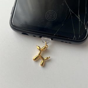 Könnte beinhalten: Ein goldfarbener Ballonhund-Anhänger an einem schwarzen Smartphone. Der Anhänger ist eine kleine, detaillierte Skulptur eines Hundes, der an der Buchse des Telefons befestigt ist. Der Bildschirm des Telefons ist gerissen.